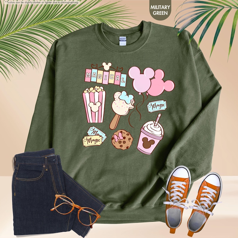 Puede incluir: Una sudadera verde con un dise&ntilde;o tem&aacute;tico de Mickey Mouse. El dise&ntilde;o incluye una pancarta que dice "MAGICAL", palomitas de ma&iacute;z, un helado de Mickey Mouse, una galleta de Mickey Mouse, un globo de Mickey Mouse y un batido de Mickey Mouse.