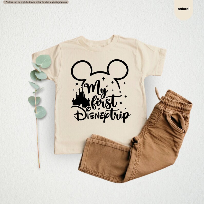 Pu&ograve; includere: Una maglietta per neonati beige con una grafica nera di orecchie di Topolino, un castello e il testo "My first Disneptrip".