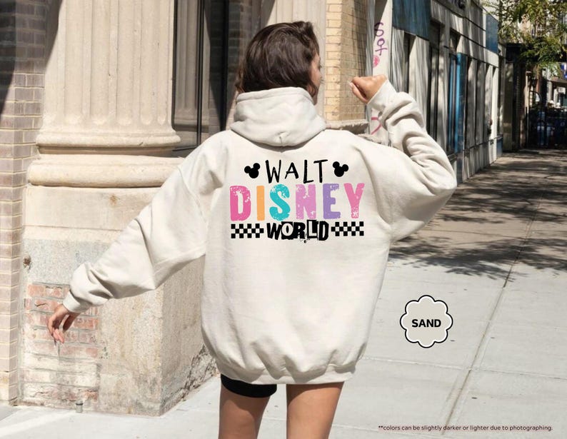 Puede incluir: Una sudadera con capucha color arena con las palabras "WALT DISNEY WORLD" en letras coloridas y desgastadas en la espalda. El texto est&aacute; acentuado con una silueta de Mickey Mouse y un patr&oacute;n de cuadros blancos y negros.