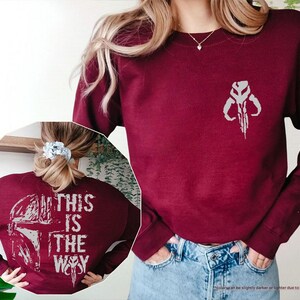 May include: A maroon sweatshirt with a white graphic of the Mandalorian symbol on the front and the text "This is the Way" on the back.