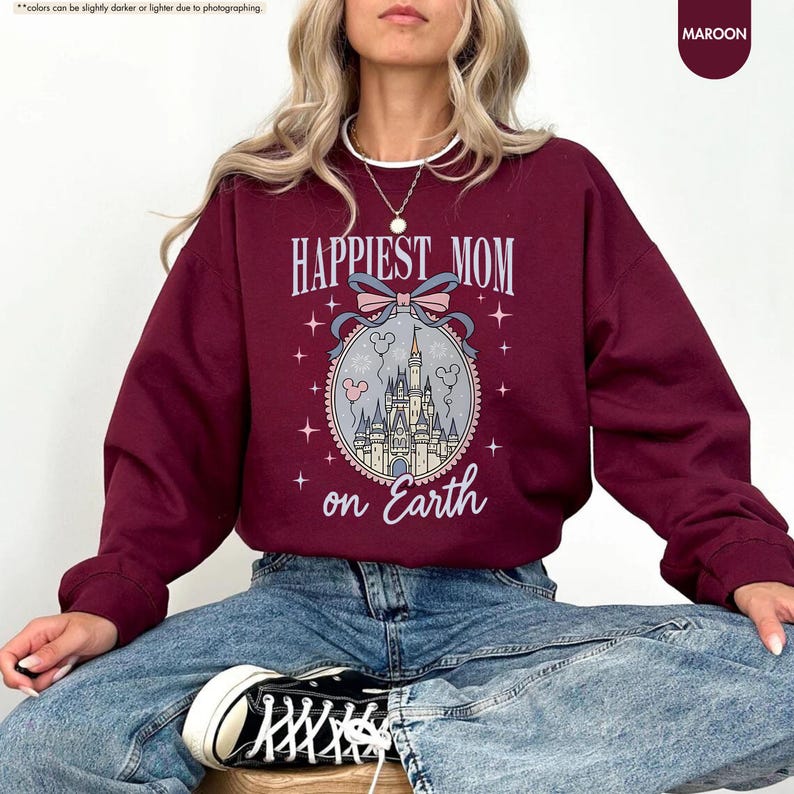 May include: Maroon sweatshirt with the text "Happiest Mom on Earth" in a decorative font. The design includes a castle illustration within an oval frame, accented with stars and a bow. The sweatshirt is a solid maroon color.