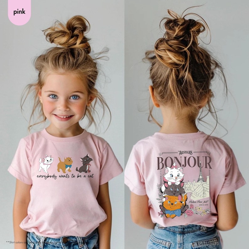 May include: Pink t-shirt featuring a cartoon cat design with the text "everybody wants to be a cat" on the front. The back of the shirt has a "Bonjour" graphic with cat illustrations and the Eiffel Tower. A young person is wearing the shirt.