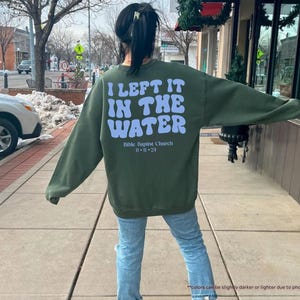May include: Military green sweatshirt with the text "I LEFT IT IN THE WATER" in light blue, along with "Bible Baptist Church" and the date "11-11-24". The sweatshirt is a long-sleeved crew neck.