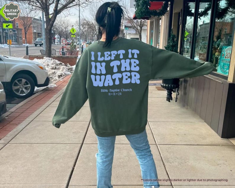 May include: Military green sweatshirt with the text "I LEFT IT IN THE WATER" in light blue, along with "Bible Baptist Church" and the date "11-11-24". The sweatshirt is a long-sleeved crew neck.