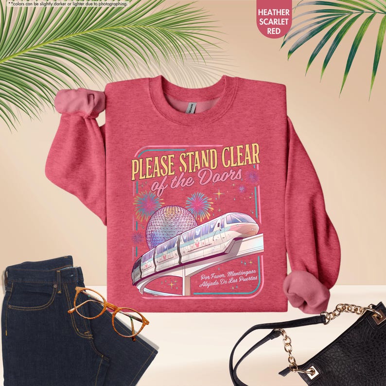 May include: Heather scarlet red sweatshirt with the text "PLEASE STAND CLEAR of the Doors" and a graphic of a monorail and fireworks. The sweatshirt is paired with blue jeans, glasses, and a black purse.