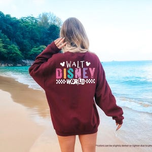 Puede incluir: Una sudadera granate con el texto "WALT DISNEY WORLD" en letras de colores. La sudadera tiene cuello redondo y mangas largas. El fondo es una escena de playa con agua azul y arena.