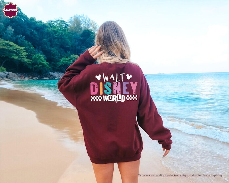 Puede incluir: Una sudadera granate con el texto "WALT DISNEY WORLD" en letras de colores. La sudadera tiene cuello redondo y mangas largas. El fondo es una escena de playa con agua azul y arena.
