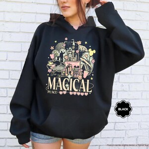 Pode incluir: Moletom preto com capuz com um gr&aacute;fico colorido com um castelo, orelhas do Mickey Mouse e o texto "The Most Magical Place". O design inclui elementos rosa, amarelo e preto. O moletom tem um bolso frontal.