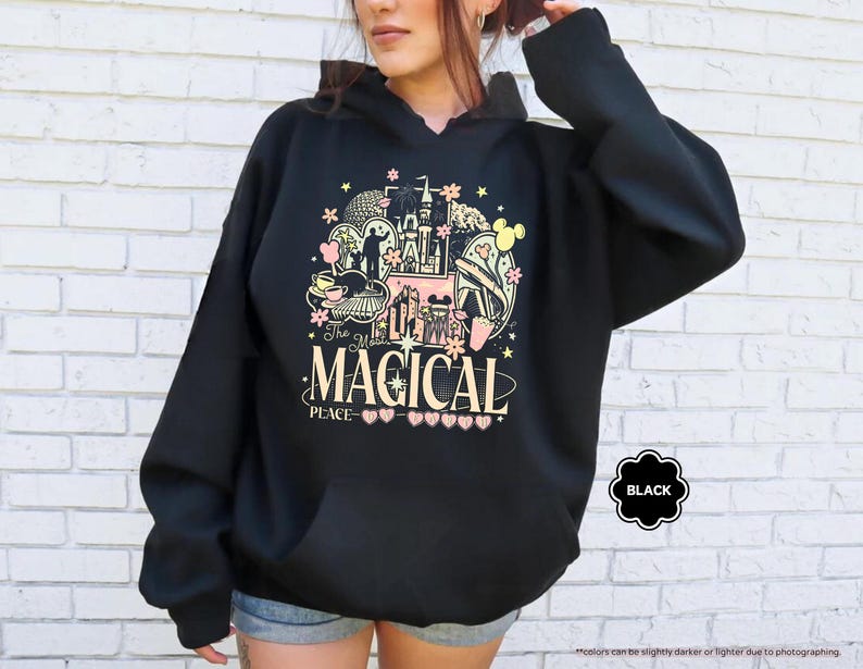 Pode incluir: Moletom preto com capuz com um gr&aacute;fico colorido com um castelo, orelhas do Mickey Mouse e o texto "The Most Magical Place". O design inclui elementos rosa, amarelo e preto. O moletom tem um bolso frontal.