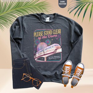 May include: Dark heather gray sweatshirt with a graphic that reads "PLEASE STAND CLEAR of the Doors" featuring a monorail and fireworks. The sweatshirt is paired with dark wash jeans and orange sneakers.