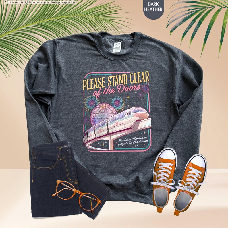 May include: Dark heather gray sweatshirt with a graphic that reads "PLEASE STAND CLEAR of the Doors" featuring a monorail and fireworks. The sweatshirt is paired with dark wash jeans and orange sneakers.