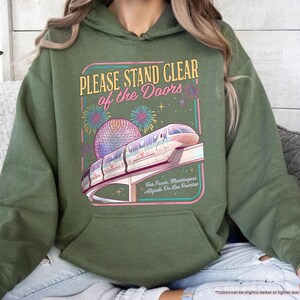 May include: Military green hoodie with a graphic that reads "PLEASE STAND CLEAR of the DOORS." The graphic features a monorail, fireworks, and a sphere. The text "Five Faur, Mantingases, Algiasts Do Ens Puertas" is also included.