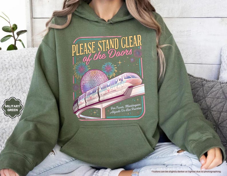 May include: Military green hoodie with a graphic that reads "PLEASE STAND CLEAR of the DOORS." The graphic features a monorail, fireworks, and a sphere. The text "Five Faur, Mantingases, Algiasts Do Ens Puertas" is also included.