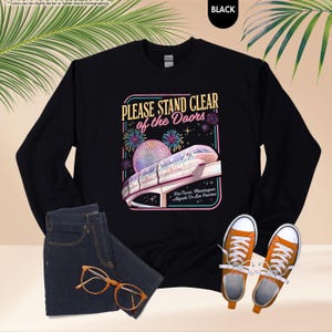 May include: Black sweatshirt with a graphic that reads "PLEASE STAND CLEAR of the Doors." The graphic features a monorail, fireworks, and a sphere. The sweatshirt is paired with blue jeans and orange sneakers.