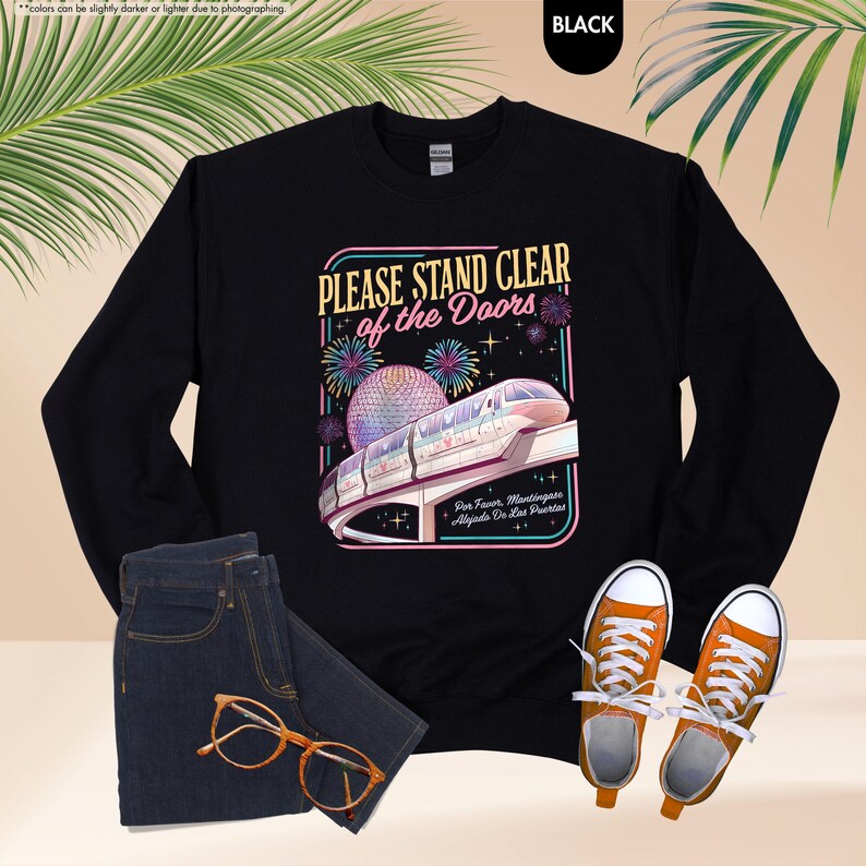 May include: Black sweatshirt with a graphic that reads "PLEASE STAND CLEAR of the Doors." The graphic features a monorail, fireworks, and a sphere. The sweatshirt is paired with blue jeans and orange sneakers.