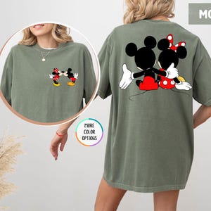 May include: A moss green t-shirt with a cartoon image of Mickey and Minnie Mouse embracing on the back. The front pocket has a smaller image of the couple kissing. The shirt has a relaxed fit.