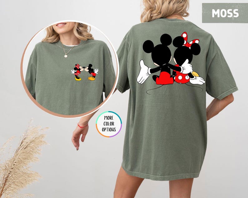 May include: A moss green t-shirt with a cartoon image of Mickey and Minnie Mouse embracing on the back. The front pocket has a smaller image of the couple kissing. The shirt has a relaxed fit.
