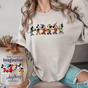 Puede incluir: Sudadera color arena con una fila de personajes de dibujos animados de Mickey Mouse. La sudadera tiene cuello redondo y mangas largas. Una imagen m&aacute;s peque&ntilde;a muestra el texto "With a Little Imagination Anything is Possible" con personajes de Mickey Mouse.