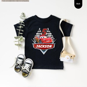 Op de afbeelding: Zwart T-shirt met een rode raceauto-afbeelding, geblokte vlaggen en het nummer 5. De tekst "JACKSON" is in rood op een witte banner gedrukt.