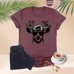 May include: A heather maroon t-shirt with a graphic of a deer wearing sunglasses and a camouflage baseball cap. The deer has large antlers.