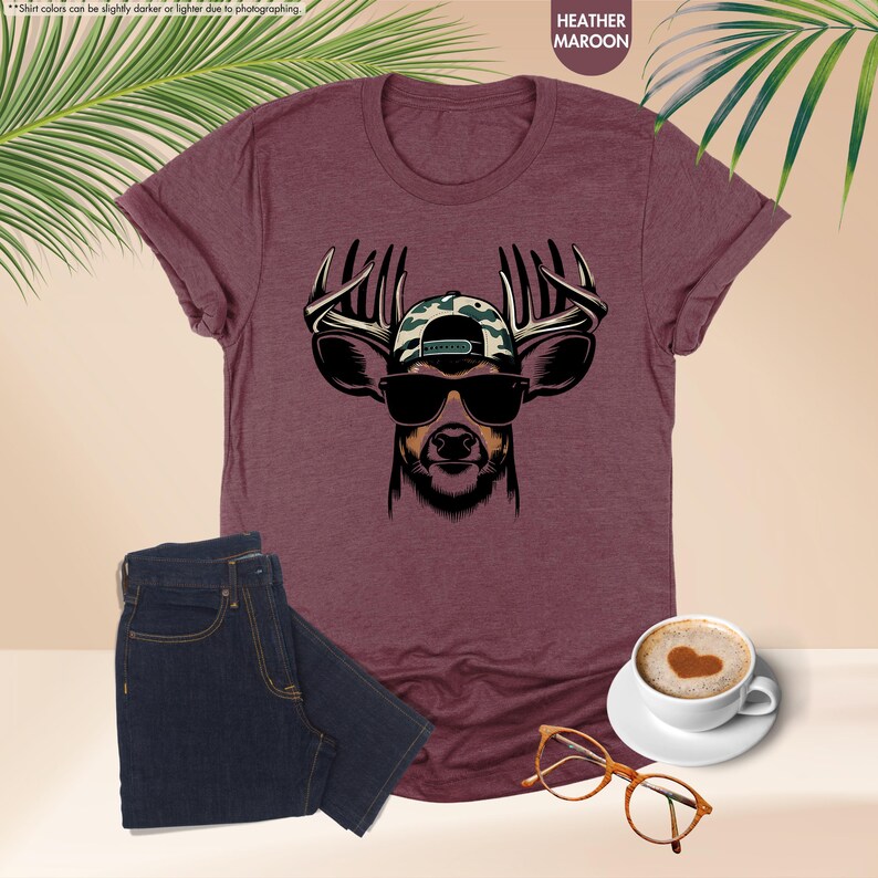 May include: A heather maroon t-shirt with a graphic of a deer wearing sunglasses and a camouflage baseball cap. The deer has large antlers.