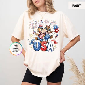 May include: Ivory-colored t-shirt featuring a patriotic graphic with two cartoon chipmunks, fireworks, and the word "USA" in red, white, and blue. The shirt has short sleeves and a relaxed fit.