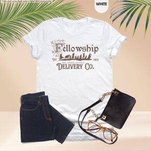 Puede incluir: Camiseta blanca con un gr&aacute;fico marr&oacute;n de los personajes de la Comunidad del Anillo caminando con el texto "Fellowship Est 1954 fast, reliable, precious DELIVERY CO."