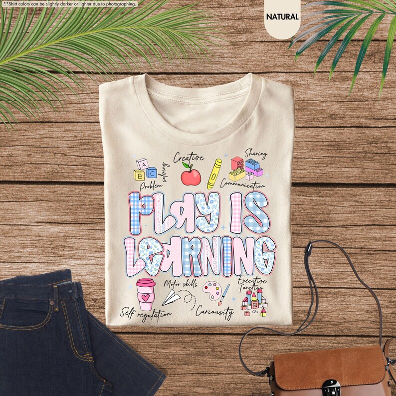 May include: A beige t-shirt with the words "PLAY IS LEARNING" in a blue and pink checkered design. The shirt has various illustrations, including an apple, pencil, blocks, and a coffee cup. The shirt is laying on a wooden surface with a pair of blue jeans.