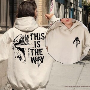 May include: A beige hoodie with a black graphic print of a Mandalorian helmet and the text "This is the Way".