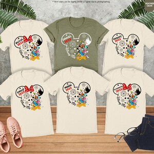May include: Six t-shirts with a Mickey Mouse ear design featuring a family of Disney characters. The shirts are in various colors, including beige, green, and white. The shirts have the text "Disney Trip" and the year "2025" printed on them.