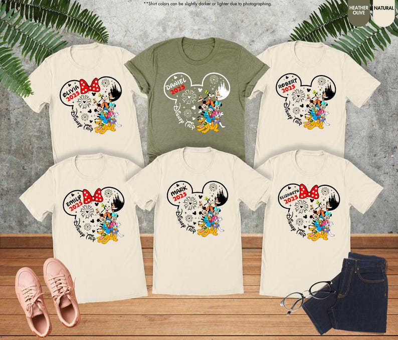 May include: Six t-shirts with a Mickey Mouse ear design featuring a family of Disney characters. The shirts are in various colors, including beige, green, and white. The shirts have the text "Disney Trip" and the year "2025" printed on them.