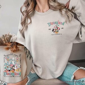 May include: A sand colored sweatshirt with a graphic print of Mickey Mouse and friends in front of the Disneyland castle. The text "Disneyland Est. 1955 WDS California" is printed on the sweatshirt.