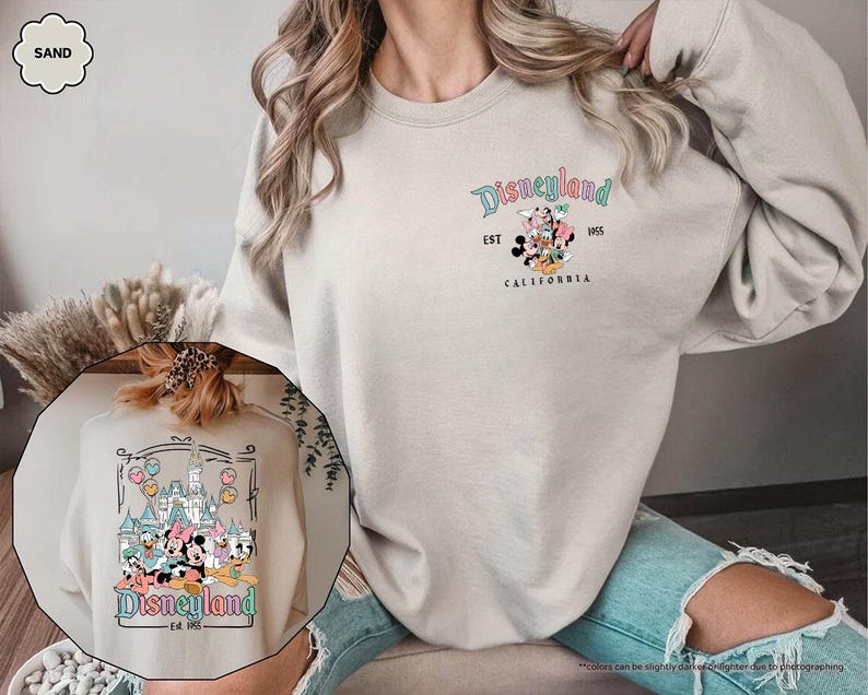 May include: A sand colored sweatshirt with a graphic print of Mickey Mouse and friends in front of the Disneyland castle. The text "Disneyland Est. 1955 WDS California" is printed on the sweatshirt.