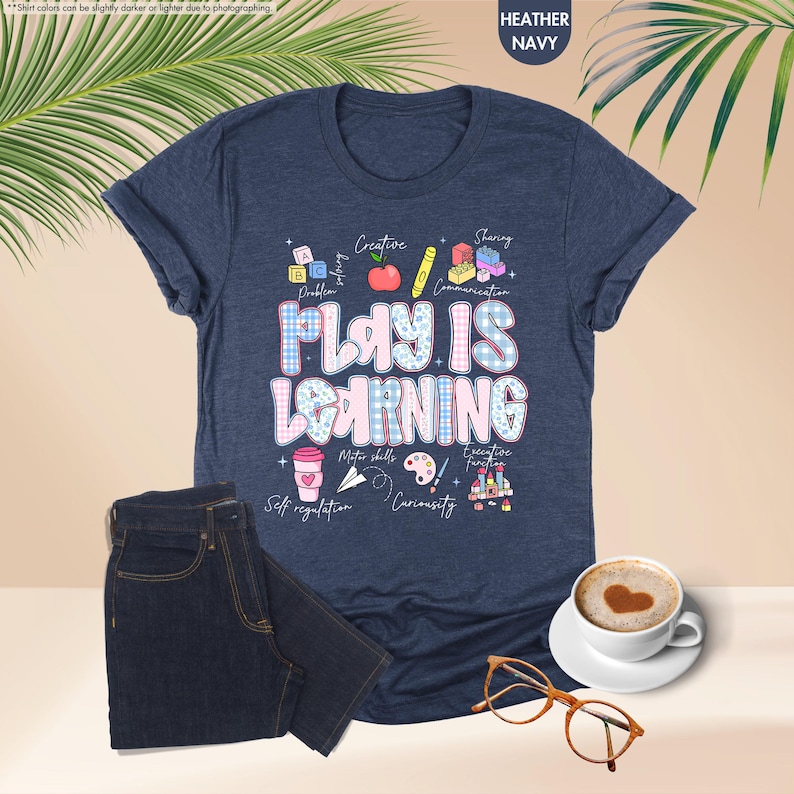 May include: Heather navy blue t-shirt with the text "PLAY IS LEARNING" in a playful font. The shirt features colorful graphics related to learning and development, including blocks, an apple, and a coffee cup. A pair of dark blue jeans is next to the shirt.