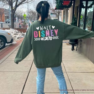 Puede incluir: Una sudadera verde militar con las palabras "WALT DISNEY WORLD" en letras de colores. El texto est&aacute; delineado en blanco y sobre un patr&oacute;n de cuadros blancos y negros. La sudadera es de manga larga.