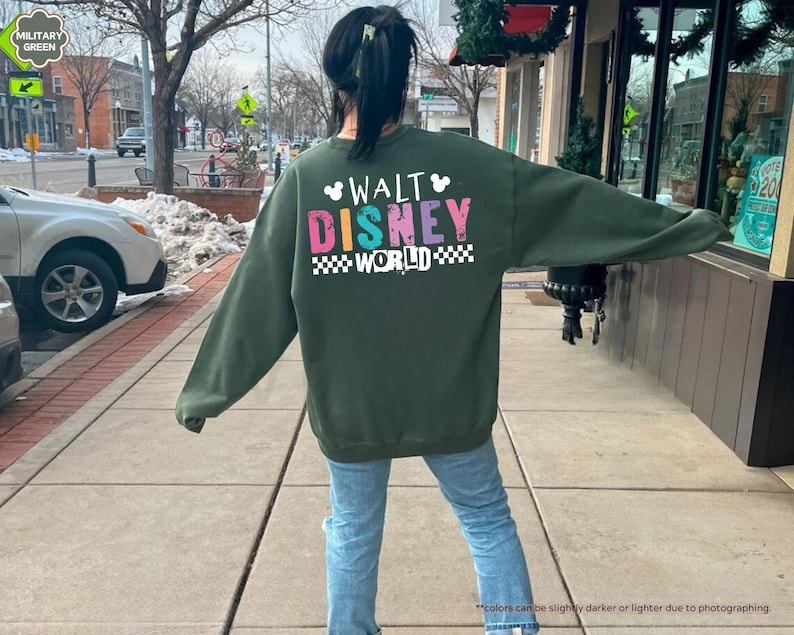 Puede incluir: Una sudadera verde militar con las palabras "WALT DISNEY WORLD" en letras de colores. El texto est&aacute; delineado en blanco y sobre un patr&oacute;n de cuadros blancos y negros. La sudadera es de manga larga.