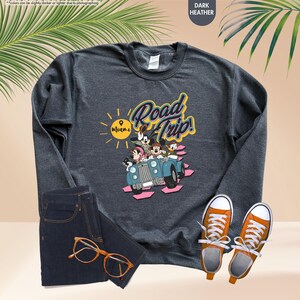 Op de afbeelding: Donkergrijs sweatshirt met een afbeelding van Mickey Mouse, Minnie Mouse, Donald Duck en Goofy in een blauwe auto met de tekst "Road Trip! Miami". Het sweatshirt wordt gecombineerd met een blauwe jeans en oranje sneakers.