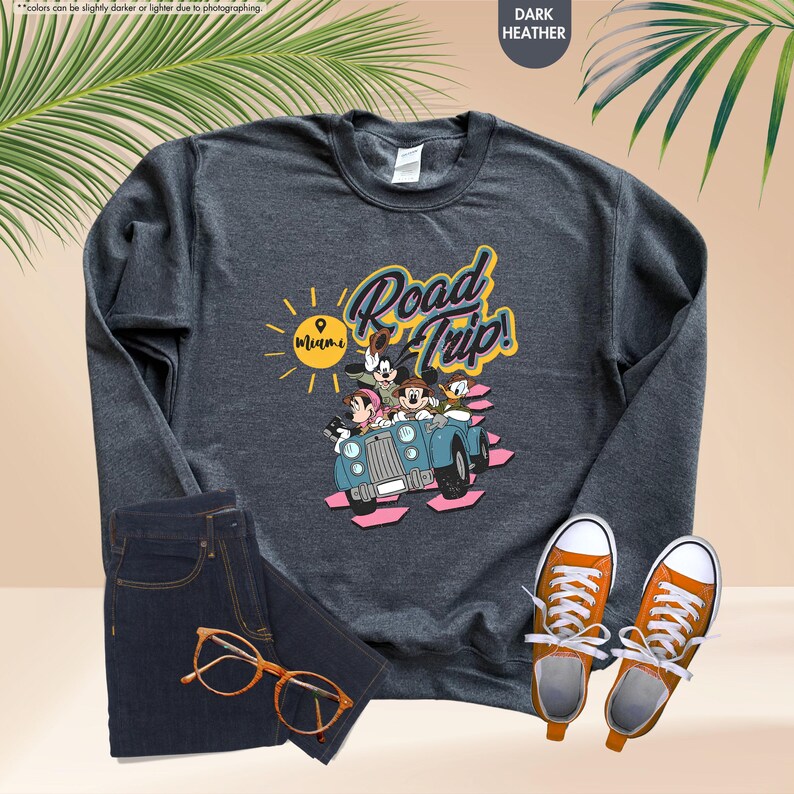 Op de afbeelding: Donkergrijs sweatshirt met een afbeelding van Mickey Mouse, Minnie Mouse, Donald Duck en Goofy in een blauwe auto met de tekst "Road Trip! Miami". Het sweatshirt wordt gecombineerd met een blauwe jeans en oranje sneakers.