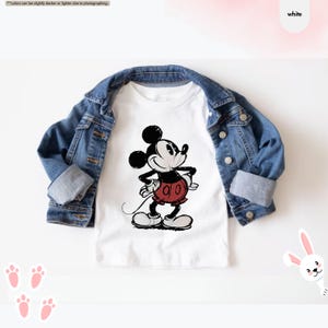 May include: A white t-shirt featuring a black and red graphic of Mickey Mouse, paired with a denim jacket. The cartoon character is depicted with a classic pose. The outfit is styled on a white background.