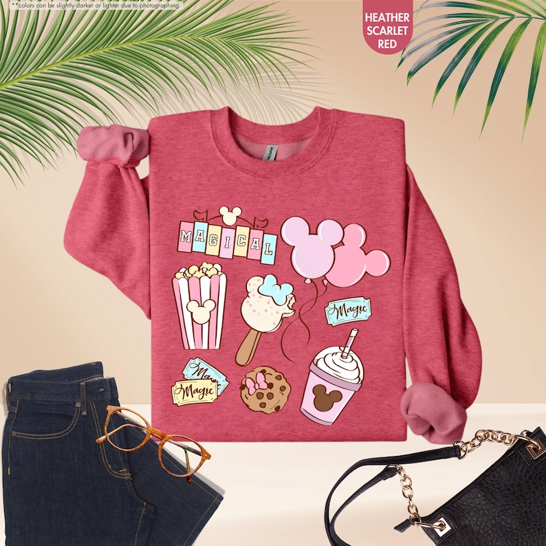 Puede incluir: Una sudadera de color rojo escarlata jaspeado con un estampado gr&aacute;fico de art&iacute;culos con tem&aacute;tica de Mickey Mouse, que incluyen palomitas de ma&iacute;z, una barra de helado de Mickey Mouse, una galleta de Mickey Mouse, un globo de Mickey Mouse y una bebida con tem&aacute;tica de Mickey Mouse. El texto "MAGICAL" est&aacute; impreso en la sudadera.