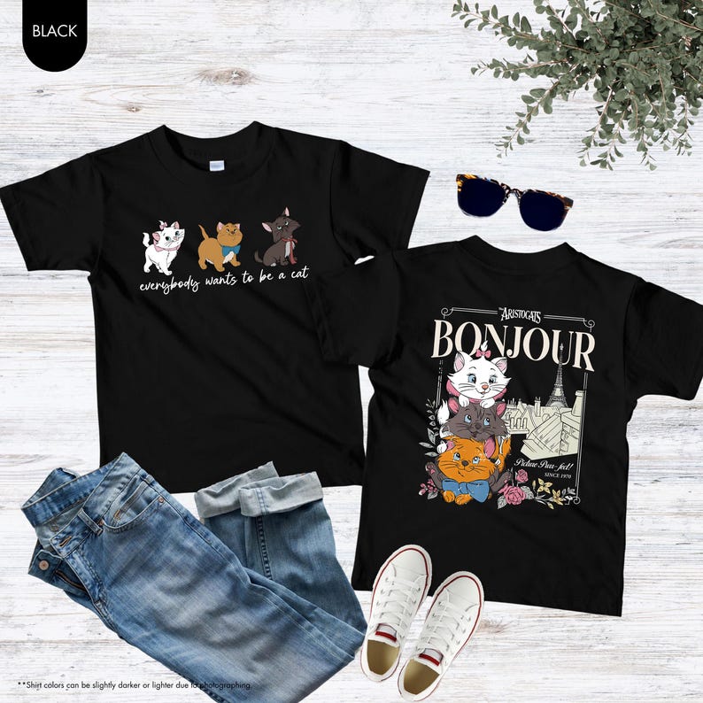 May include: Two black t-shirts with cat graphics and the text "everybody wants to be a cat" and "Bonjour". The shirts are paired with blue jeans and white sneakers. Sunglasses and a plant are also in the image.