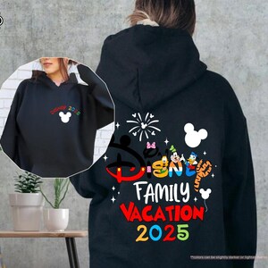 K&ouml;nnte beinhalten: Schwarzer Kapuzenpullover mit einem bunten Disney Family Vacation 2025-Motiv mit Mickey Mouse und anderen Disney-Charakteren. Das Motiv enth&auml;lt auch Feuerwerk und eine Mickey-Mouse-Ohrensilhouette.