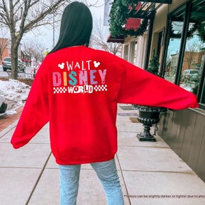 Puede incluir: Una sudadera roja brillante con las palabras "WALT DISNEY WORLD" en letras de bloque coloridas. El texto est&aacute; en la parte posterior de la sudadera. La sudadera tiene mangas largas y cuello redondo.