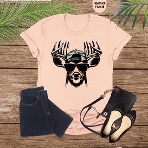 May include: A pink t-shirt with a graphic of a deer wearing sunglasses and a camouflage baseball cap. The deer has antlers and a black nose.