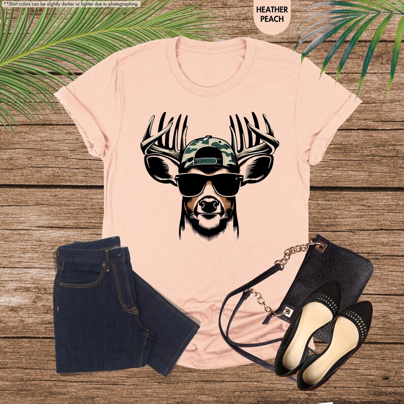May include: A pink t-shirt with a graphic of a deer wearing sunglasses and a camouflage baseball cap. The deer has antlers and a black nose.