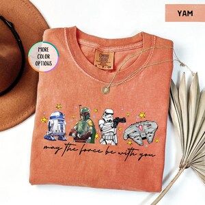 Puede incluir: Una camiseta de manga larga color &ntilde;ame con los personajes de Star Wars R2-D2, Boba Fett, un Stormtrooper y el Halc&oacute;n Milenario. La frase "may the force be with you" est&aacute; escrita debajo. Un collar de oro est&aacute; sobre la camiseta.
