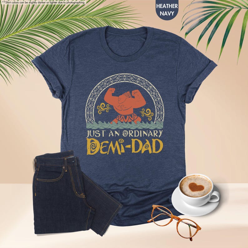 Puede incluir: Una camiseta azul marino jaspeada con una imagen de dibujos animados de un hombre musculoso con un cuerpo rojo y un taparrabos amarillo. El hombre est&aacute; de pie con los brazos levantados por encima de su cabeza. El texto "JUST AN ORDINARY DEMI-DAD" est&aacute; impreso debajo de la imagen.
