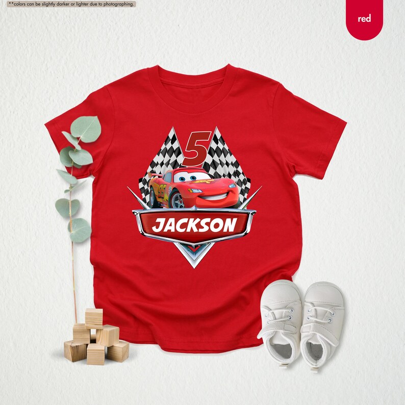 Op de afbeelding: Rood T-shirt met een afbeelding van een rode raceauto met nummer 5 en de naam JACKSON erop gedrukt. De auto is omringd door een geruit vlagontwerp.