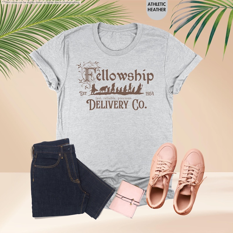 Puede incluir: Una camiseta gris jaspeada con un gr&aacute;fico marr&oacute;n de los personajes de la Comunidad del Anillo caminando en fila. El texto "Fellowship" est&aacute; por encima de los personajes y "EST 1954 fast, reliable, precious DELIVERY CO." est&aacute; por debajo de los personajes.