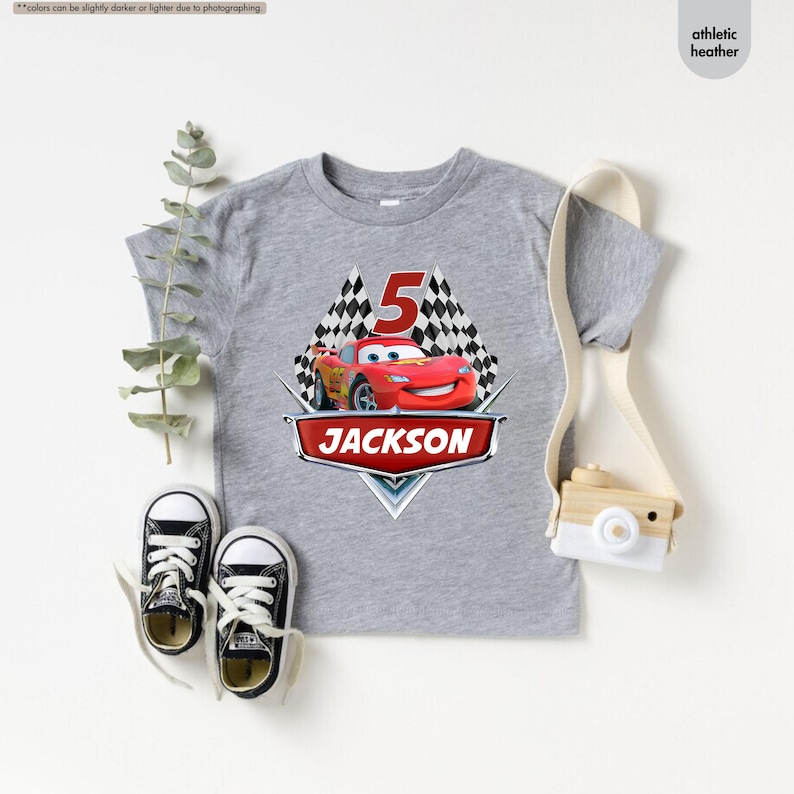 Op de afbeelding: Grijs T-shirt met korte mouwen met een rode raceauto-afbeelding, geblokte vlaggen en het nummer 5. De tekst "JACKSON" is onder de auto gedrukt.